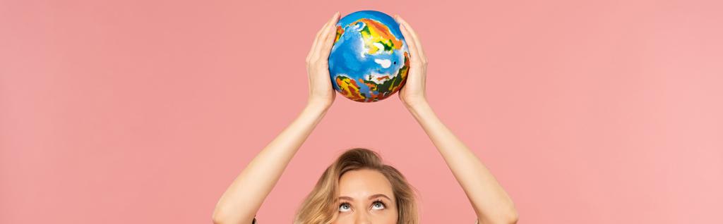 Blonde woman holding globe above head isolated on pink, panoramic shot - 写真・画像