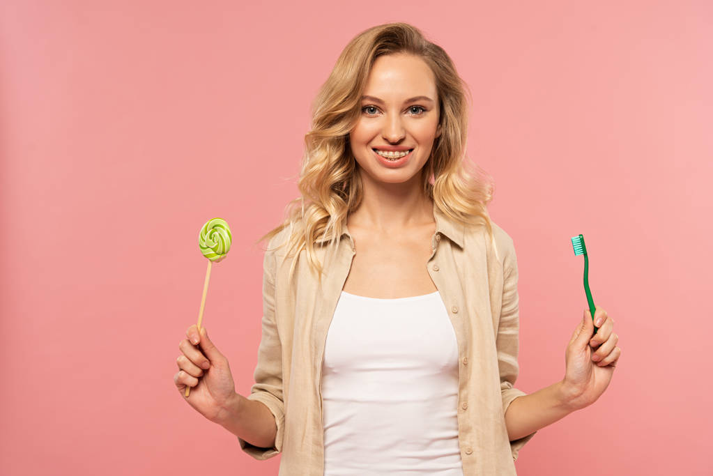 Smiling blonde woman holding toothbrush and lollipop isolated on pink - 写真・画像