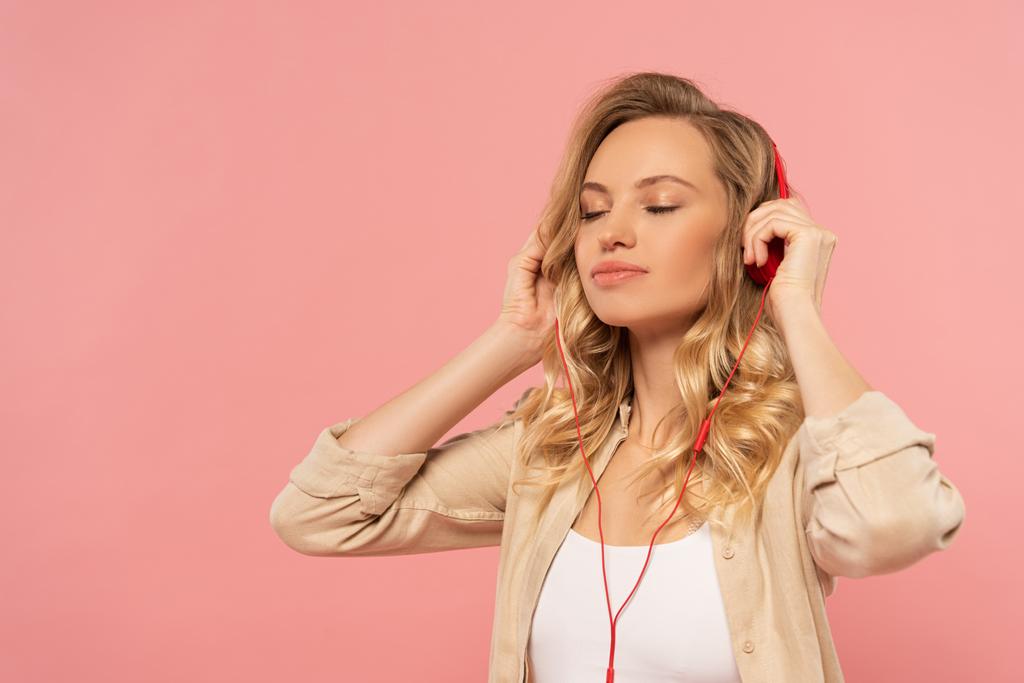 Blonde woman with closed eyes in headphone isolated on pink - 写真・画像