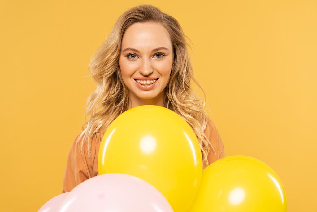 Mujer joven sonriente sosteniendo globos de colores aislados en amarillo
 - Foto, imagen