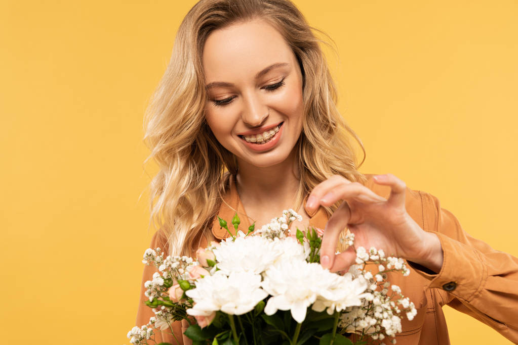 Smiling woman touching flower in bouquet isolated on yellow - 写真・画像