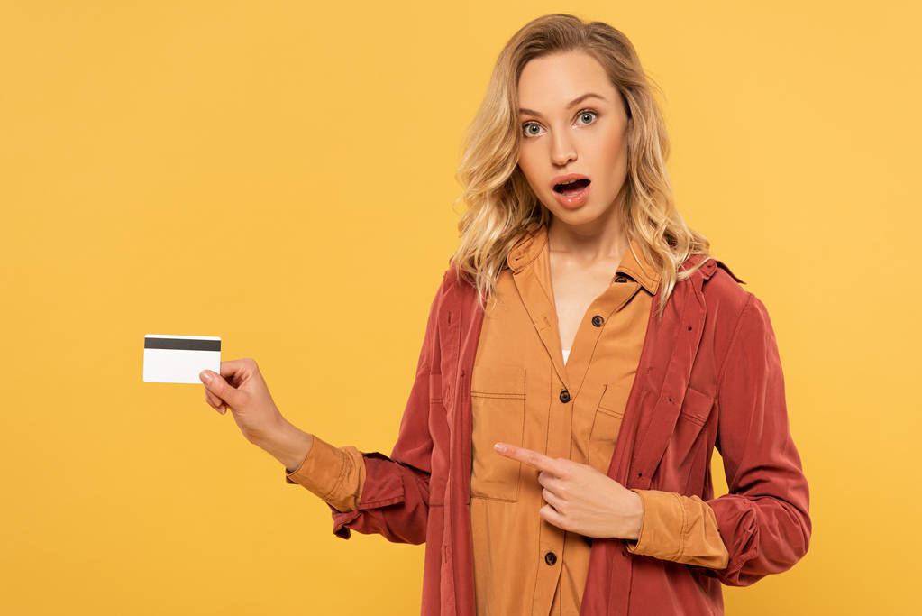 Järkyttynyt blondi nainen osoittaa luottokortilla eristetty keltainen
 - Valokuva, kuva
