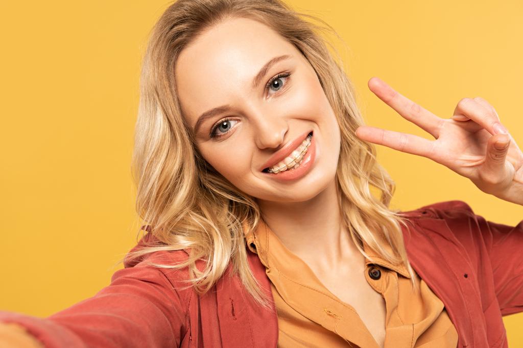 Улыбающаяся женщина в зубных скобках показывает знак мира, делая селфи, изолированное на желтом
 - Фото, изображение