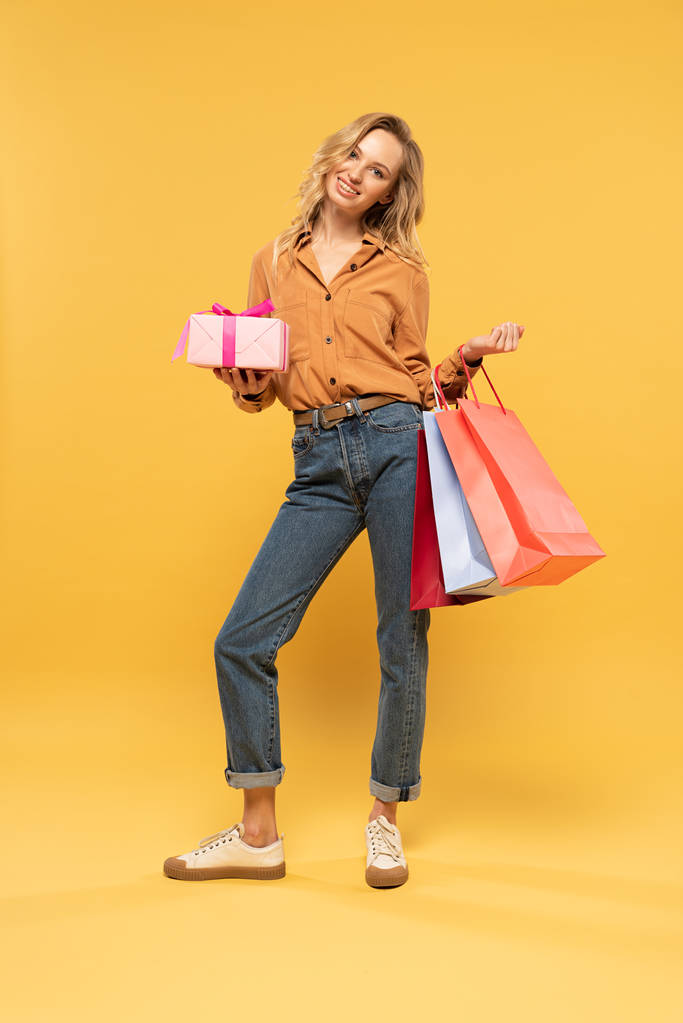 Lächelnde Frau mit Geschenkbox und Einkaufstaschen auf gelbem Hintergrund - Foto, Bild