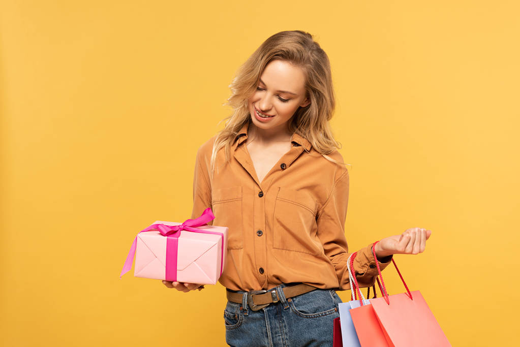 Smiling woman looking at gift box and holding shopping bags isolated on yellow - 写真・画像