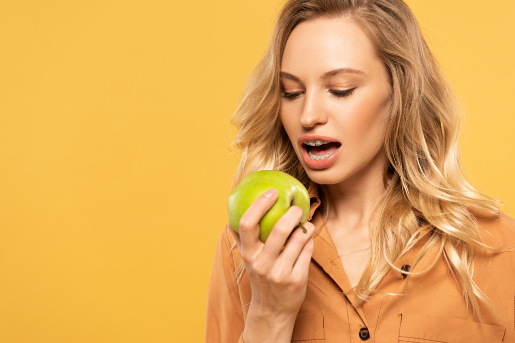 Young woman with dental braces biting green apple isolated on yellow - Photo, Image
