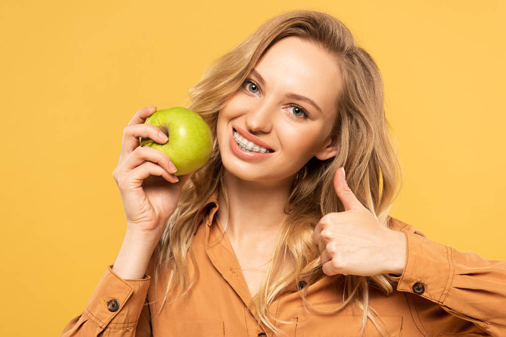 Smiling woman holding green apple and showing thumb up gesture isolated on yellow - Photo, Image