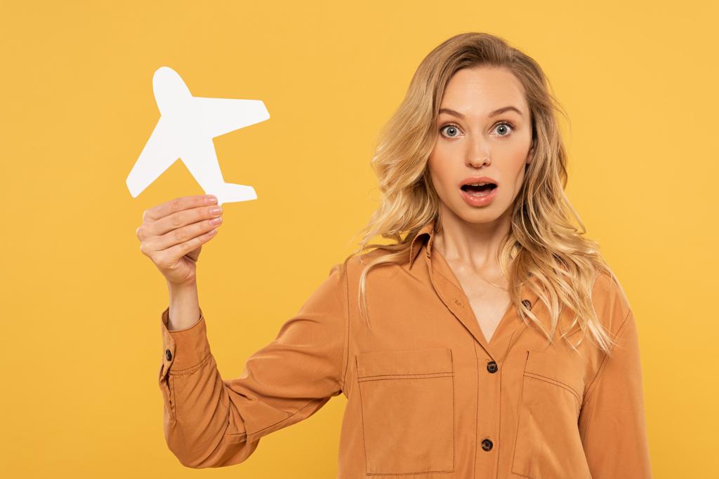 Surprised blonde woman holding paper plane isolated on yellow - 写真・画像