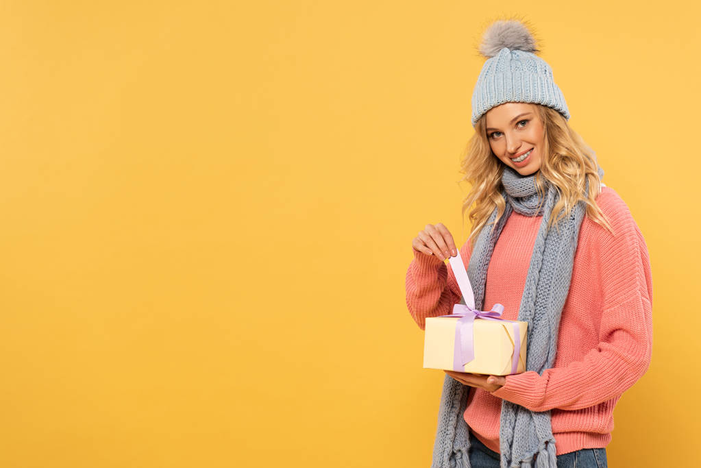 Lächelnde Frau mit Hut und Schal hält Schleife der Geschenkbox isoliert auf gelb - Foto, Bild