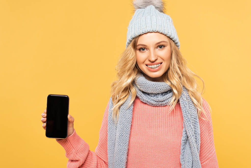 Smiling woman in hat and scarf holding smartphone with blank screen isolated on yellow - 写真・画像
