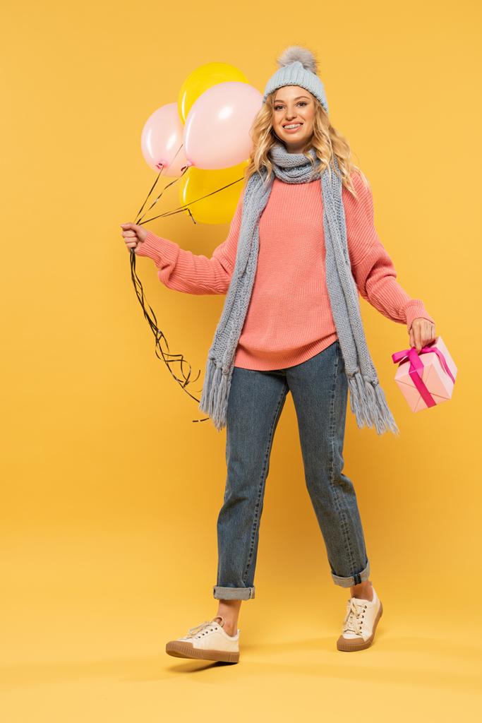 Şapkalı ve atkılı gülümseyen kadın elinde balonlar ve sarı arka planda hediye kutusu tutuyor. - Fotoğraf, Görsel