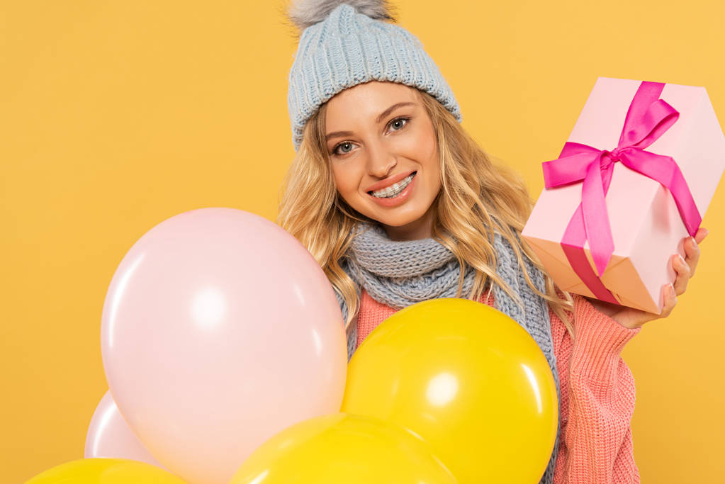 усміхнена жінка в капелюсі і шарфі тримає повітряні кулі і подарункову коробку ізольовано на жовтому
 - Фото, зображення