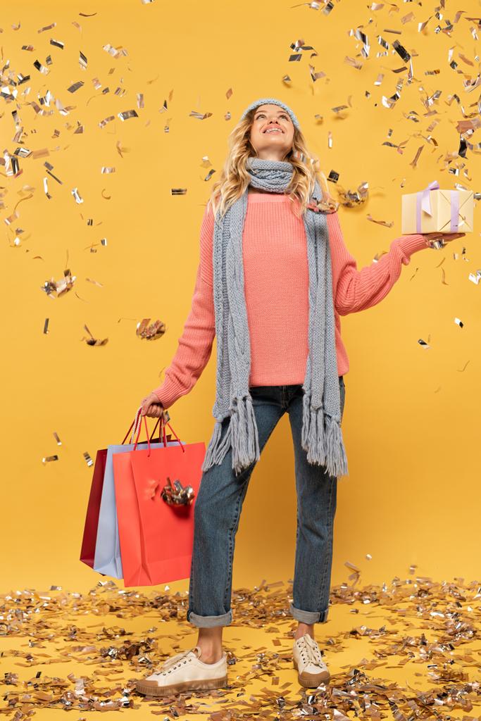 Smiling woman in hat holding gift box and shopping bags looking at golden confetti isolated on yellow background - 写真・画像