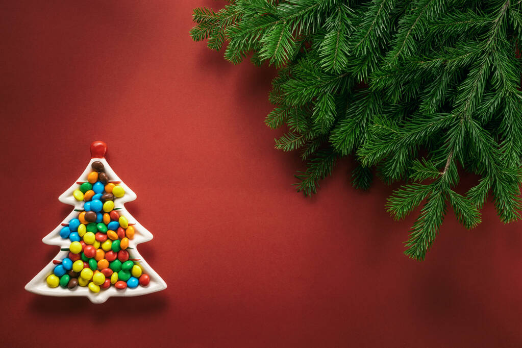 Composizione natalizia. Un piatto a forma di albero di Natale pieno di caramelle colorate. Rami di abete rosso su fondo rosso. Layout piatto, vista dall'alto, spazio di copia
. - Foto, immagini