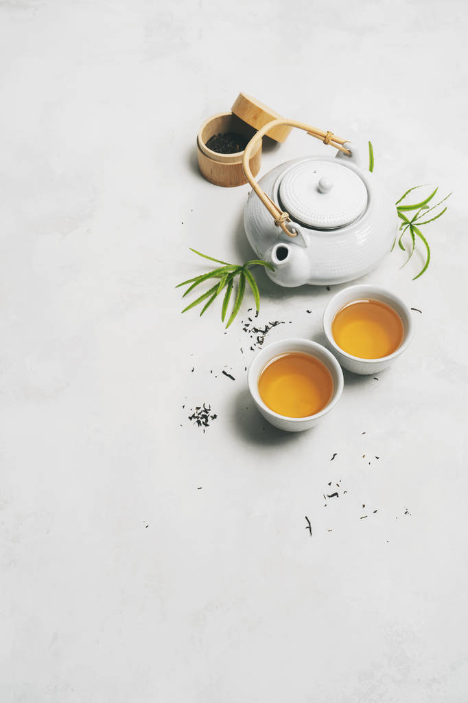 Aziatisch thee concept, twee witte kopjes thee, theepot, Theeset, eetstokjes, bamboe mat omgeven door droge groene thee op witte achtergrond met ruimte voor tekst. Brouw- en drinkthee. - Foto, afbeelding
