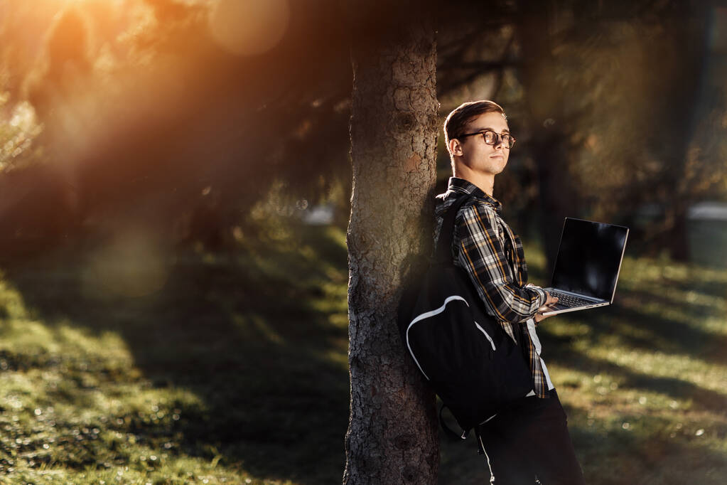 Όμορφος νεαρός φοιτητής σε περιστασιακή στολή κοιτάξουμε μακριά, ενώ στέκεται μπροστά από το δέντρο με σημειωματάριο ή φορητό υπολογιστή στο πάρκο. - Φωτογραφία, εικόνα
