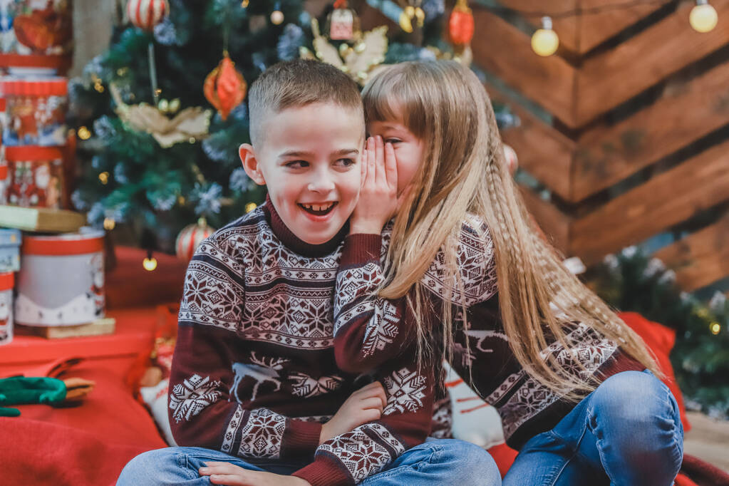 Sweet shot van een meisje vertellen een geheim in het oor van haar broer zitten op rode deken thuis, celbrating Kerstmis. - Foto, afbeelding