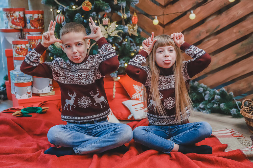 Crianças sorridentes em blusas de lã mostram orelhas grandes com as mãos e põem línguas no fundo de madeira com árvore de natal.
. - Foto, Imagem