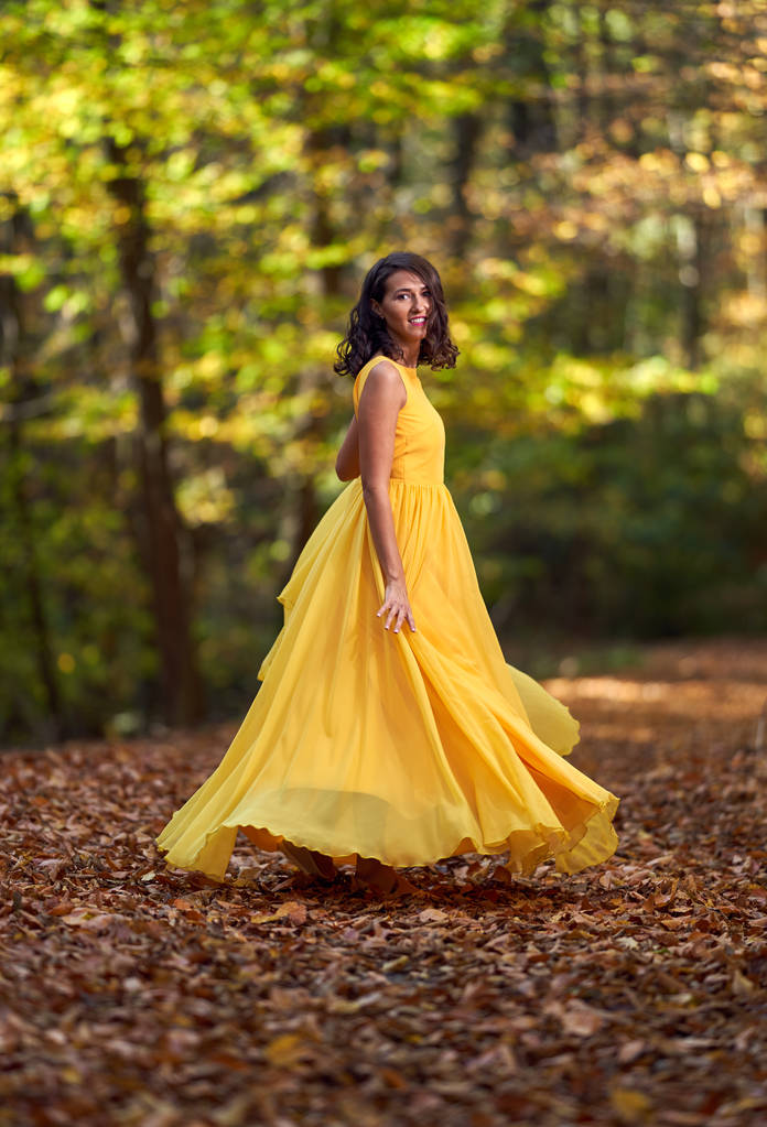 Boldog fiatal nő hosszú sárga ruhában táncol egy őszi tájon - Fotó, kép