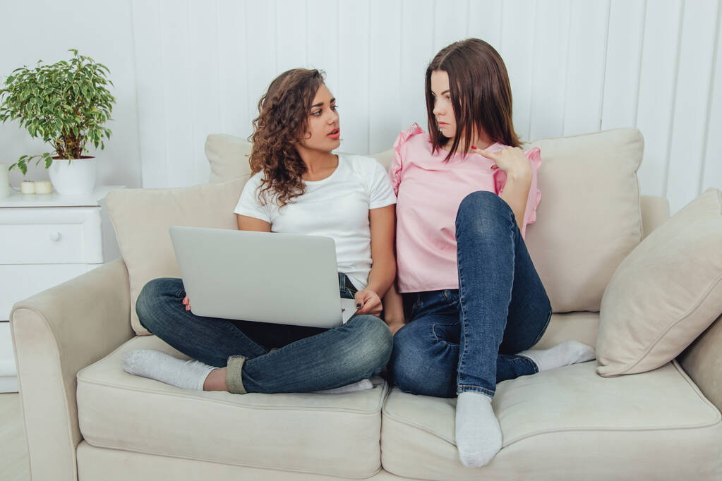 Дві милі дівчата сидять на дивані з ноутбуком, серфінгом в Інтернеті, розмовляють. Їхні ноги схрещені
. - Фото, зображення