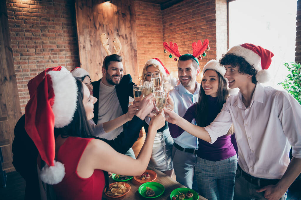 Photo de réunion de groupe fête de Noël de nombreuses personnes célébrant clinking verres à vin boisson vin mousseux vêtements formalwear santa chapeaux cornes de cerf debout autour de la table à l'intérieur
 - Photo, image