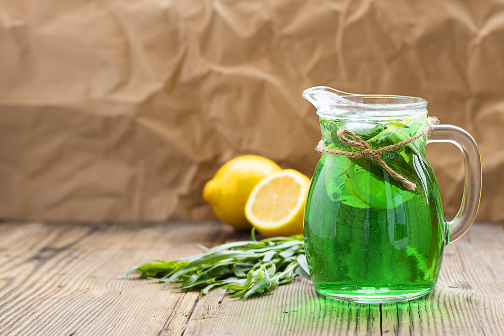 Letní studený zelený nápoj z estragonu, s mátou a plátky citronu ve skleněné karafě v ohnisku a zelenými listy estragonu a citrónem rozostřeným vzadu. Šumivý nápoj. Vodorovně s mezerou pro kopírování - Fotografie, Obrázek