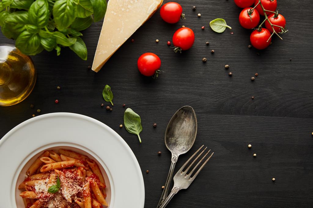 vrchní pohled na chutné boloňské těstoviny s rajčatovou omáčkou a parmezánem v bílém talíři v blízkosti ingrediencí a příborů na černém dřevěném pozadí - Fotografie, Obrázek