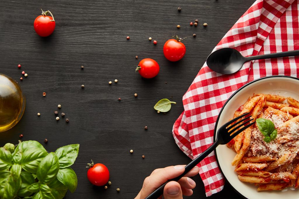abgeschnittene Ansicht des Mannes, der leckere Bolognese-Pasta mit Tomatensauce und Parmesan von weißem Teller auf schwarzem Holztisch mit Zutaten isst und Serviette überprüft - Foto, Bild