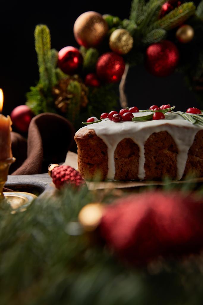 selective focus of traditional Christmas cake with cranberry near Christmas wreath with baubles on wooden table isolated on black - Photo, Image