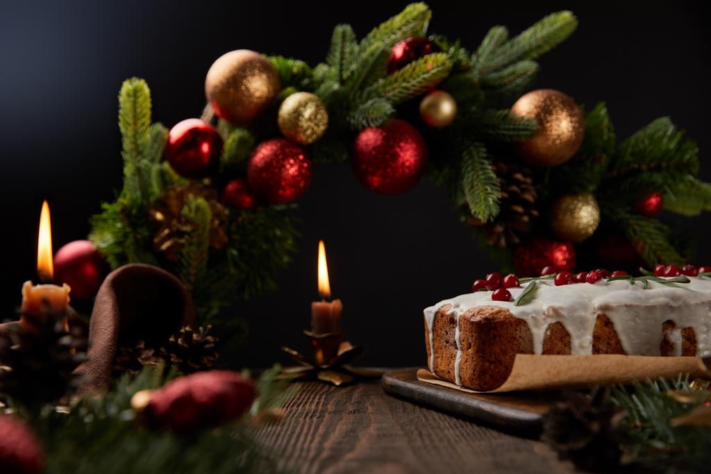 traditional Christmas cake with cranberry near Christmas wreath with baubles and burning candles on wooden table isolated on black - Photo, Image