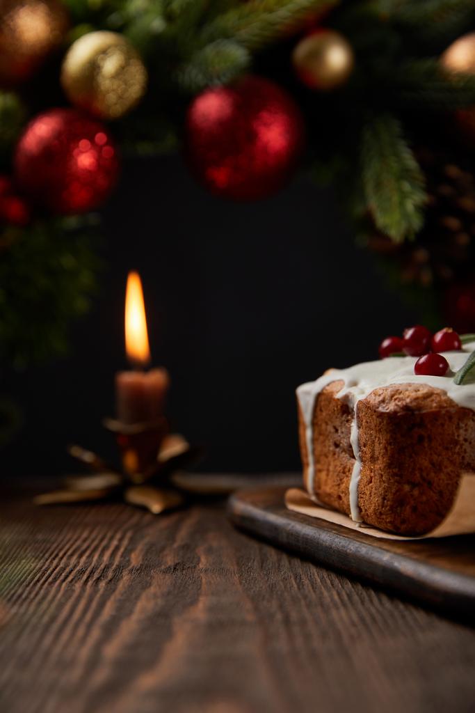 foco selectivo de pastel tradicional de Navidad con arándano cerca de la corona de Navidad con adornos y vela encendida en la mesa de madera aislado en negro
 - Foto, Imagen