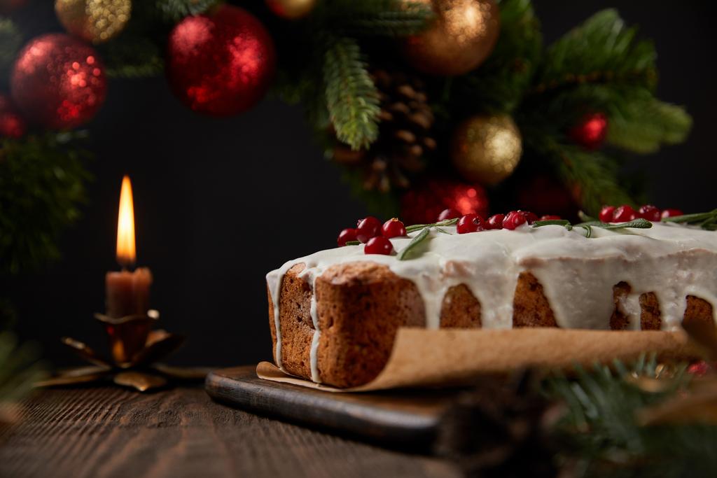 selektywne skupienie tradycyjnego tortu bożonarodzeniowego z żurawiną w pobliżu świątecznego wieńca z bombkami i świecą na drewnianym stole odizolowanym na czarno - Zdjęcie, obraz