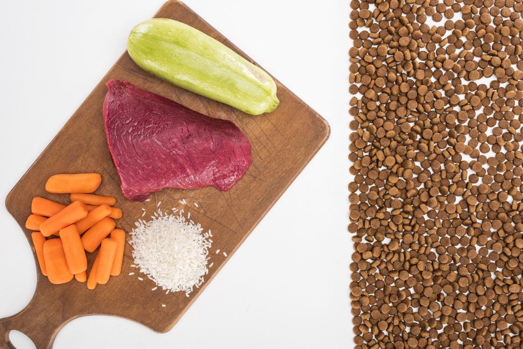 vrchní pohled na suché krmivo pro zvířata v blízkosti syrového masa, rýže, mrkve a cukety na dřevěné řezací desce izolované na bílém - Fotografie, Obrázek