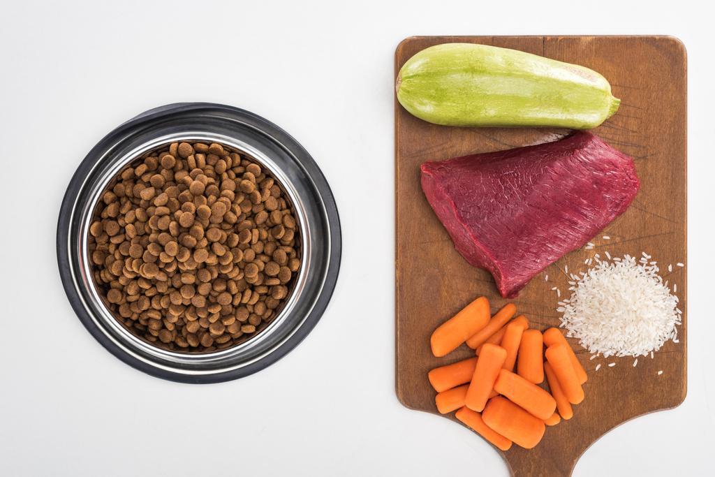 top view of dry pet food and raw meat, rice, carrot and zucchini on wooden cutting board isolated on white - 写真・画像