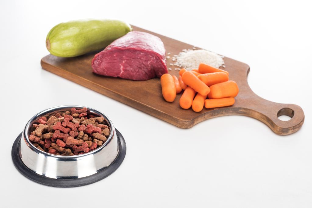 comida seca para mascotas en un tazón cerca de carne cruda, arroz, zanahoria y calabacín en una tabla de cortar de madera aislada en blanco
 - Foto, Imagen