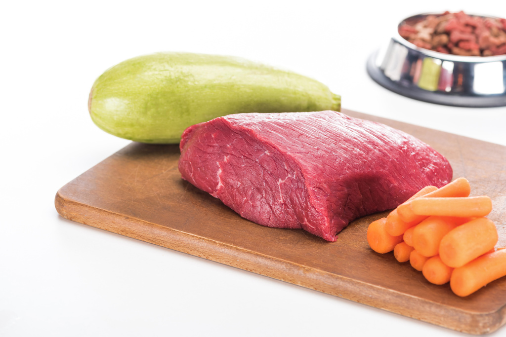 selectieve focus van droog voeder voor gezelschapsdieren in de buurt van rauw vlees, wortel en courgette op houten snijplank geïsoleerd op wit - Foto, afbeelding