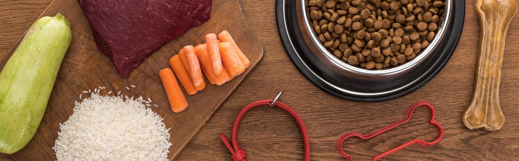 top view of dry pet food in silver bowl near raw meat, carrot, rice, zucchini, bone, collar and pet toy on wooden table, panoramic shot - Photo, Image
