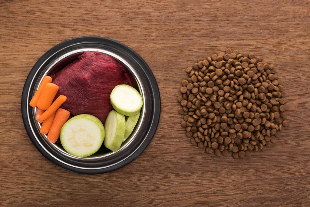 vista superior de alimentos secos para mascotas cerca de verduras crudas y carne en un tazón sobre una mesa de madera
 - Foto, Imagen