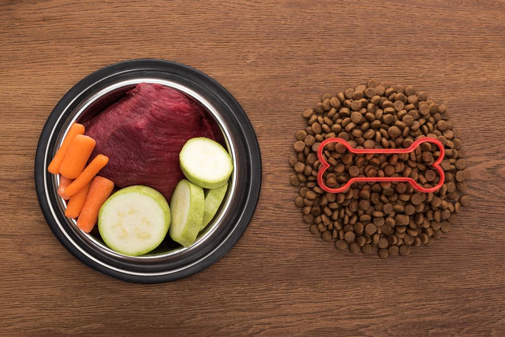 верхний вид сухого корма для домашних животных с костью рядом с сырыми овощами и мясом в миске на деревянном столе
 - Фото, изображение
