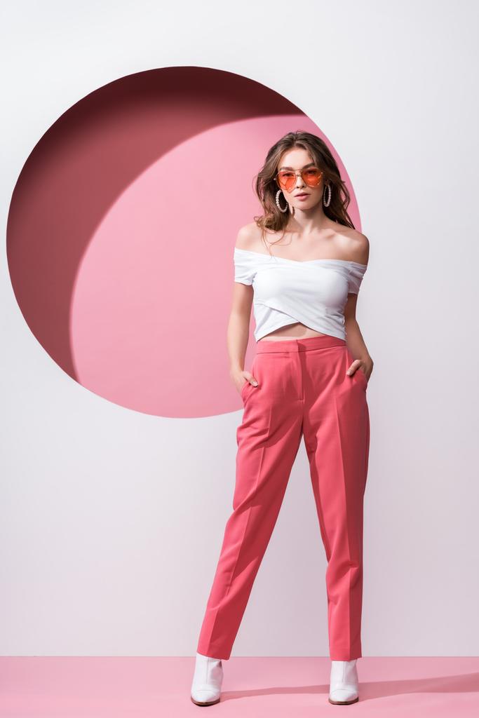 attractive girl in sunglasses standing with hands in pockets on pink and white  - Photo, Image