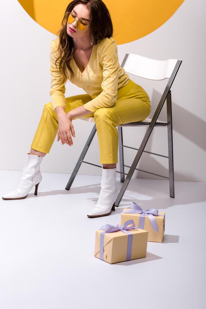 ελκυστική κοπέλα με γυαλιά ηλίου κάθεται στην καρέκλα κοντά δώρα σε πορτοκαλί και άσπρο  - Φωτογραφία, εικόνα