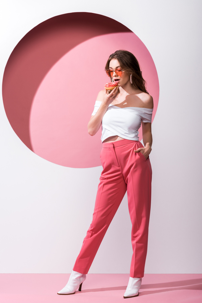 beautiful girl holding tasty doughnut and standing with hand in pocket on pink and white  - 写真・画像