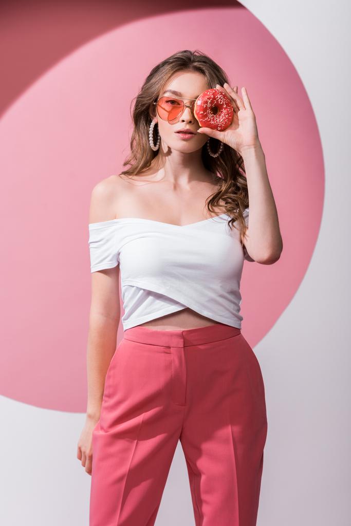 όμορφη κοπέλα που καλύπτει το πρόσωπο με νόστιμο ντόνατ και στέκεται σε ροζ και λευκό  - Φωτογραφία, εικόνα
