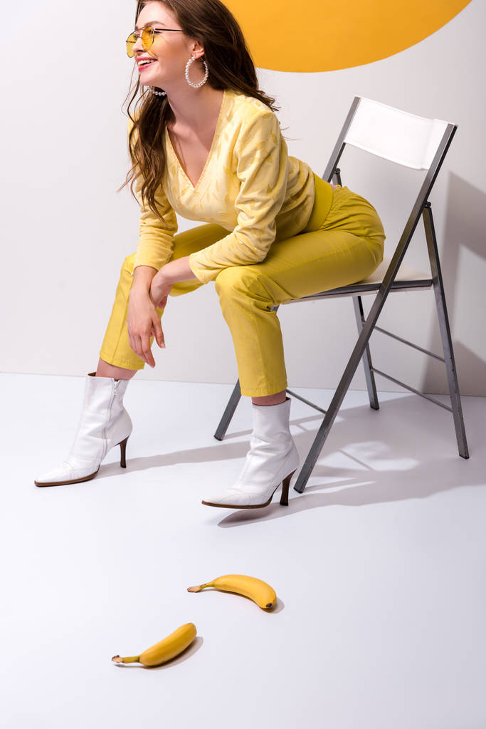 Ευτυχισμένη γυναίκα χαμογελώντας ενώ κάθεται στην καρέκλα κοντά σε κίτρινες μπανάνες σε λευκό  - Φωτογραφία, εικόνα
