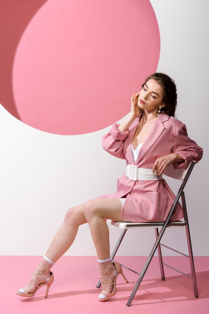 stylish woman in blazer with belt sitting on chair on white and pink  - 写真・画像