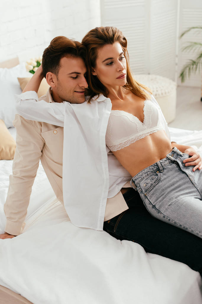 girlfriend in bra hugging and sitting on smiling boyfriend in apartment  - Photo, Image