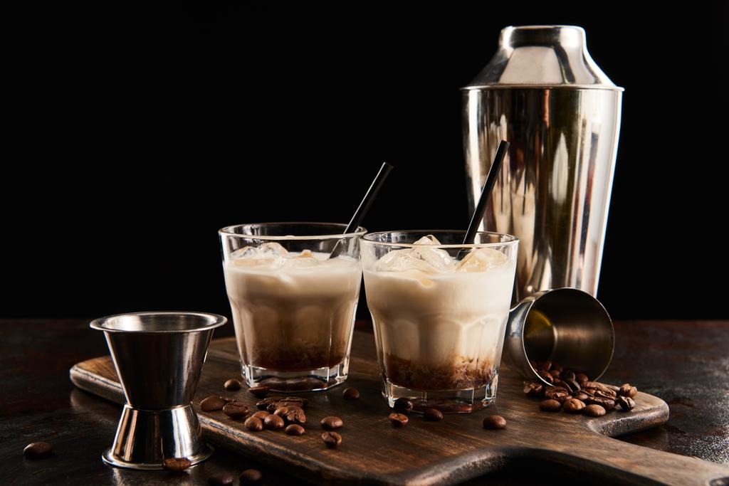 white russian cocktail in glasses with straws on wooden board with coffee grains and shaker isolated on black - Photo, Image