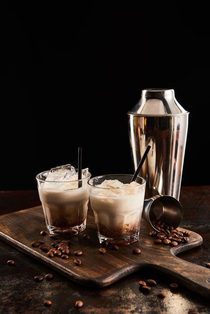 white russian cocktail in glasses with straws on wooden board with coffee grains and shaker isolated on black - Photo, Image