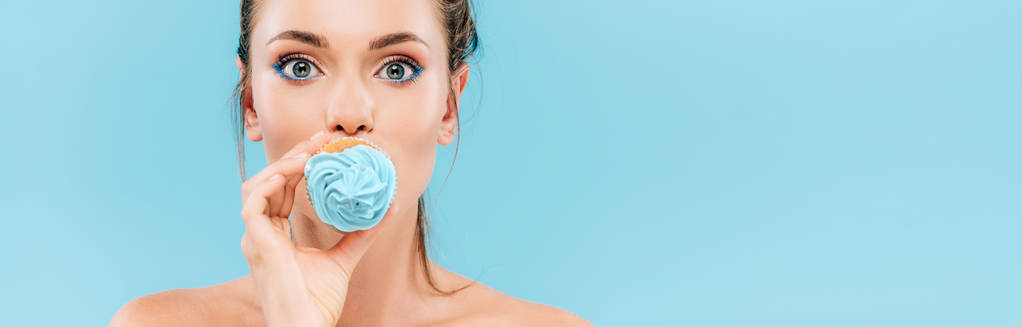 nahá krásná žena držící cupcake na ústech izolované na modré, panoramatický výstřel - Fotografie, Obrázek