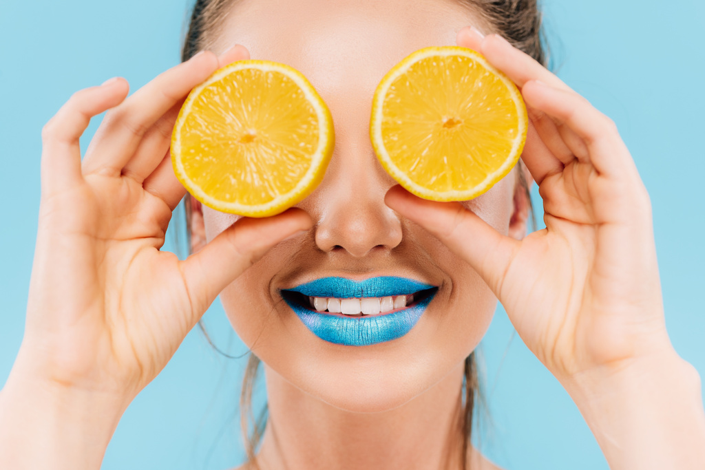 sonriente hermosa mujer con labios azules sosteniendo mitades anaranjadas en los ojos aislados en azul
 - Foto, imagen